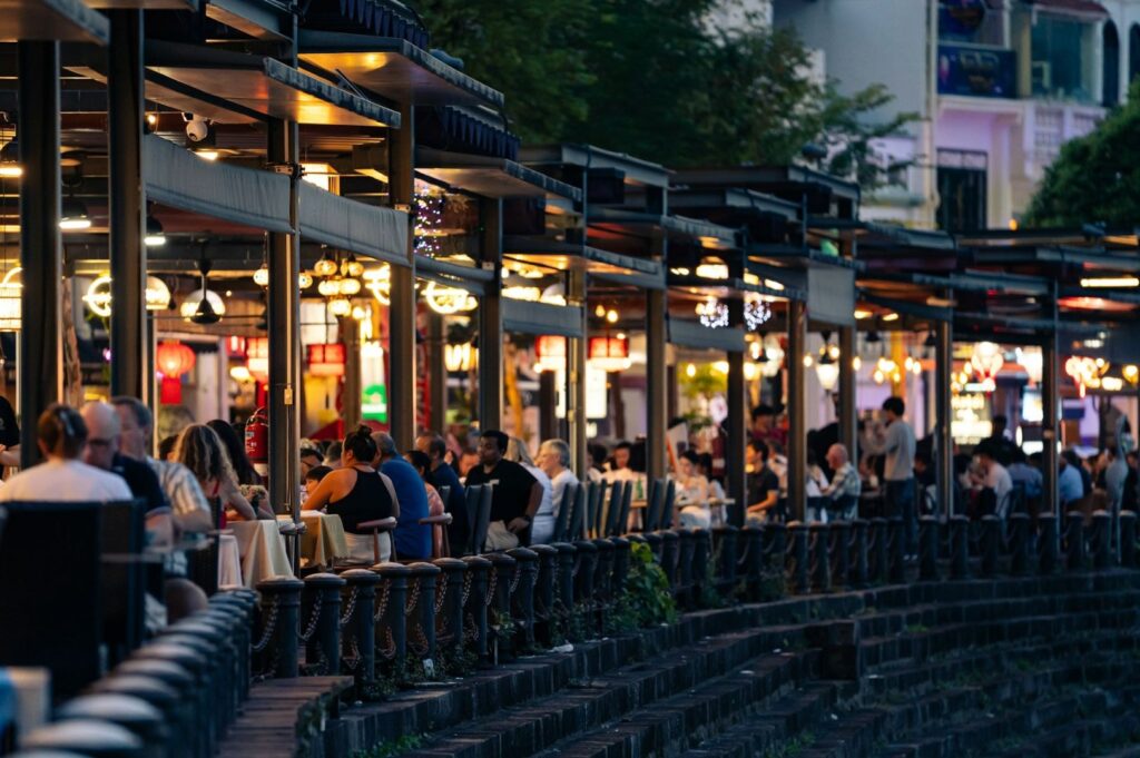 Outdoor restaurant with warmly lit dining areas and seated patrons enjoying a meal. The atmosphere is lively and social, under a twilight sky.