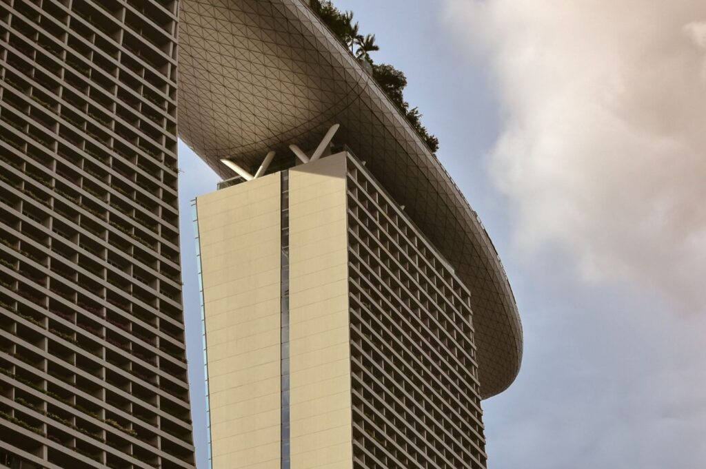 A modern high-rise with a unique rooftop garden shaped like a boat spans across multiple towers under a partly cloudy sky, conveying innovation and grandeur.