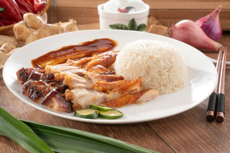 A plate of Hainanese chicken rice with sliced roasted chicken, fluffy white rice, cucumber slices, and dark sauce. Background includes ginger, shallot, and chopsticks, evoking a savory and comforting meal.