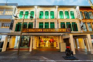 Facade of the Chinatown Heritage Centre, featuring a traditional building with green shutters, ornate detailing, and warm lighting. A bronze statue sits in front.