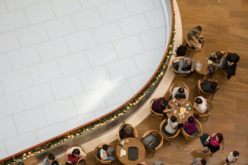 Overhead view of a café with people seated at round tables, adjacent to a large, tiled floor. Festive garlands line the railing, creating a cozy atmosphere.