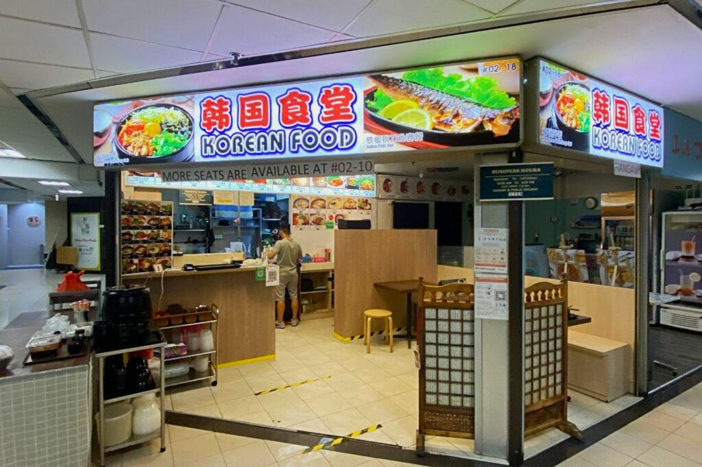 A brightly lit Korean food stall in a food court, featuring vibrant signage with images of food. A customer stands at the counter. Cozy and inviting atmosphere.