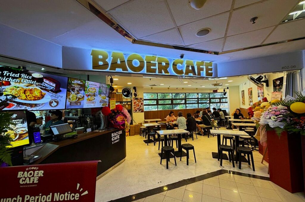 Bright café interior with a glowing "Baoer Café" sign. Customers sit at tables, and colorful menu boards display dishes. The atmosphere is lively and inviting.
