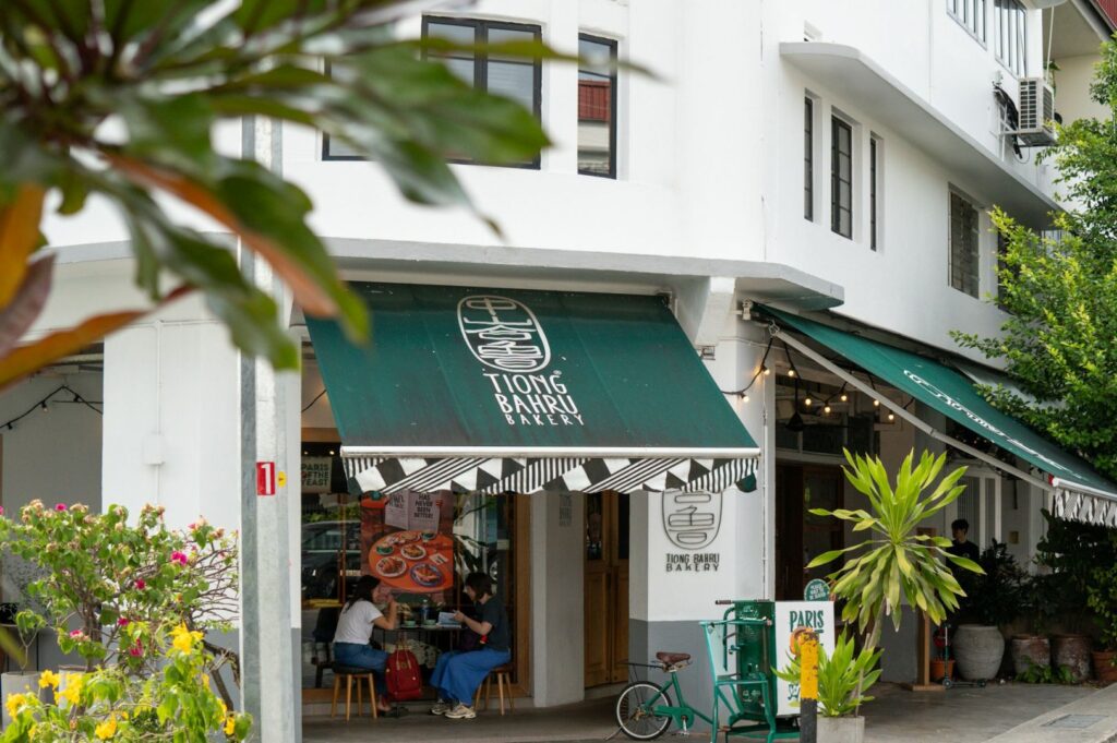 Street view of a bakery with a green awning on a corner building. Two people sit outside, surrounded by plants, creating a cozy, inviting atmosphere.