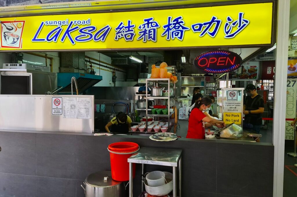 Food stall with a bright yellow sign reading "Sungei Road Laksa" and "Open" sign. Workers are preparing bowls of laksa in a clean, bustling kitchen.