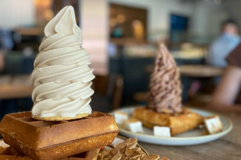 A soft serve vanilla ice cream swirl atop a waffle, garnished with almond slices. In the background, chocolate ice cream on a waffle is slightly blurred.