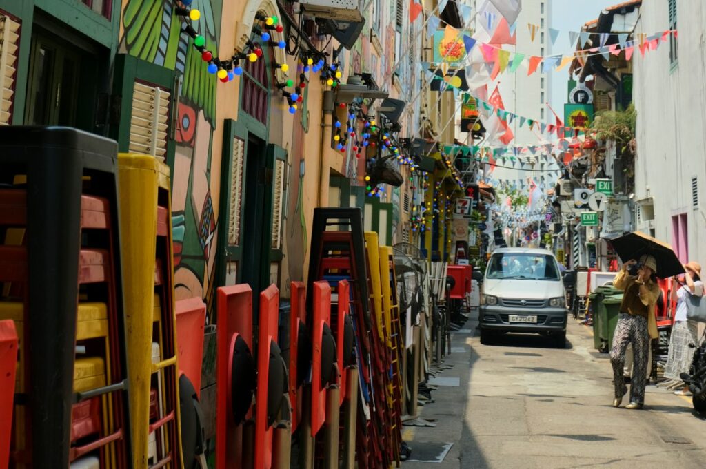 Narrow vibrant alley with colorful flags, murals, and garlands. A car passes pedestrians holding umbrellas, evoking a festive, lively atmosphere.