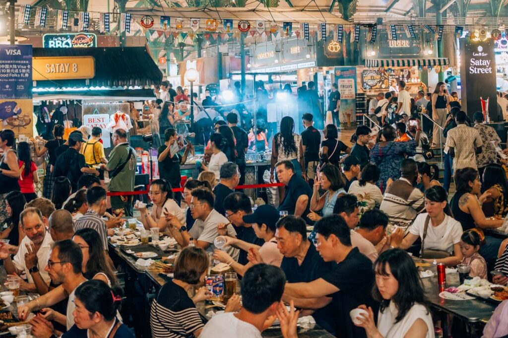 Busy night market with diverse crowd enjoying food at communal tables. Colorful stalls and festive banners create lively, vibrant atmosphere.