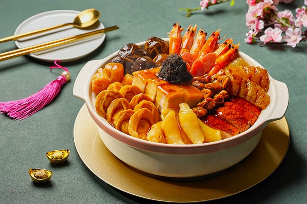 A vibrant dish of Poon Choi featuring shrimp, mushrooms, tofu, and abalone is artfully arranged in a pot. Gold ingots and flowers add festive decor.
