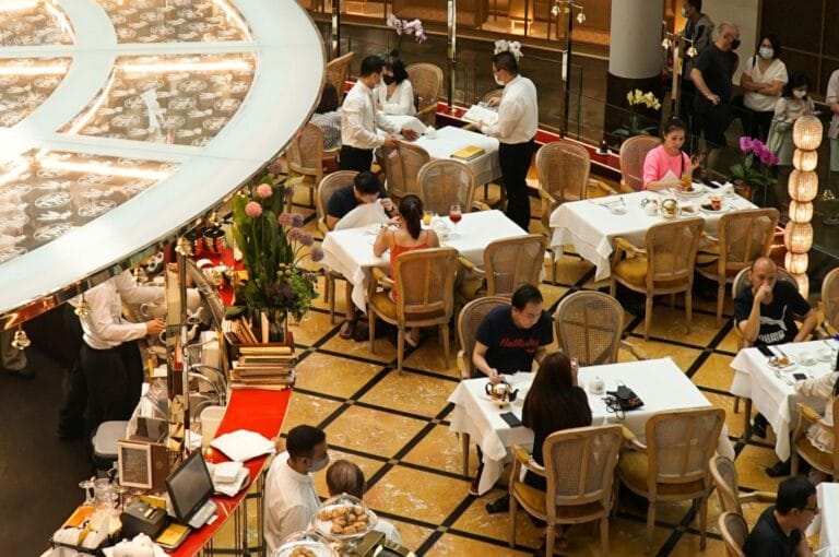 A bustling restaurant scene with diners at white-clothed tables, waitstaff taking orders. Warm lighting, flower arrangements, and casual ambiance create a lively atmosphere.