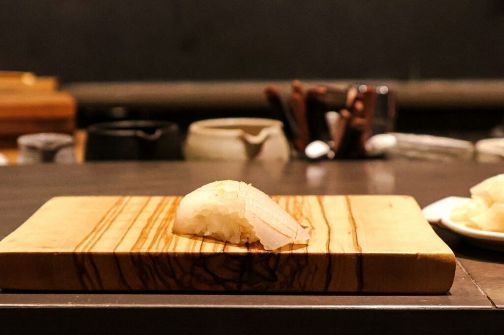 Sushi on a wooden board in a dimly lit restaurant. The board shows wood grain patterns, with small bowls and cutlery blurred in the background, creating a refined atmosphere.