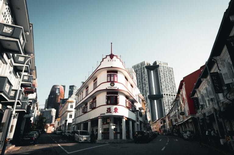 Street scene in Singapore featuring a historic white building with red accents and the year “1939” displayed. Modern skyscrapers loom in the background, creating a contrast between old and new architecture under a clear sky.