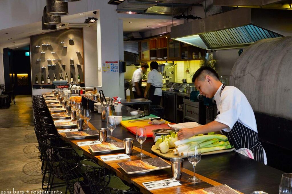 Modern kitchen with chefs. One chef prepares vegetables on a counter covered with neatly set tables and glasses, creating a busy yet inviting atmosphere.