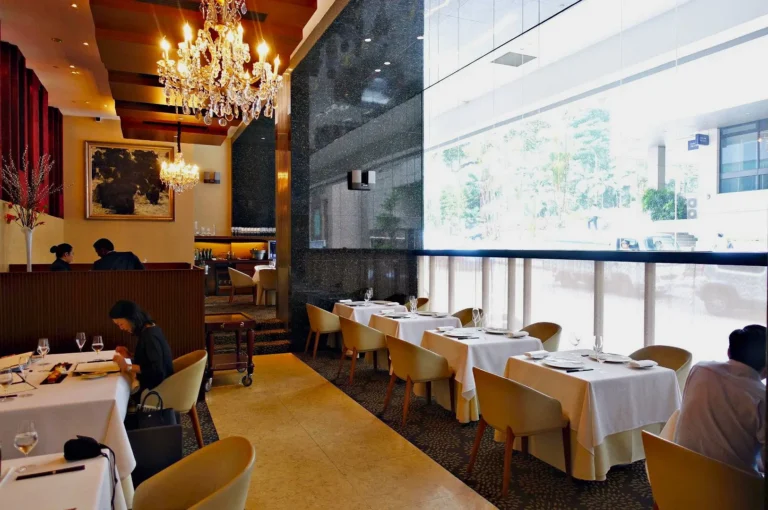Elegant restaurant interior with chandeliers, large windows, and white tablecloths. Diners sit at tables, creating a refined and serene ambiance.