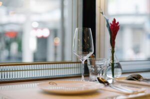 Elegant restaurant table setting with a wine glass, water glass, and vase with a red flower on a white tablecloth. Soft, natural light from a window.