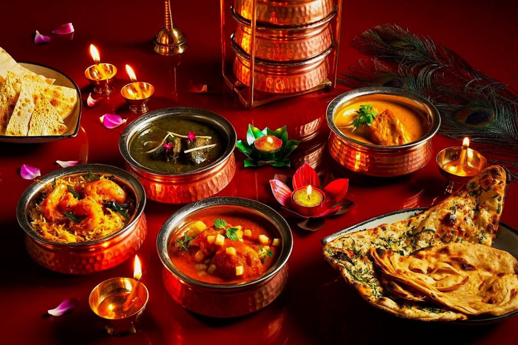 An assortment of Indian dishes in copper bowls on a red table, highlighted by glowing candles and rose petals. The scene is warm and festive.
