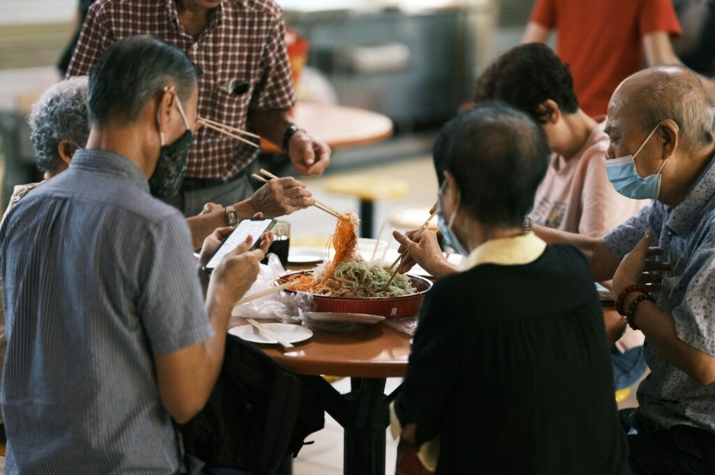 A group of people gathers around a table, joyfully using chopsticks to serve a dish of noodles and vegetables. One person wears a mask, adding a sense of caution to the social atmosphere.