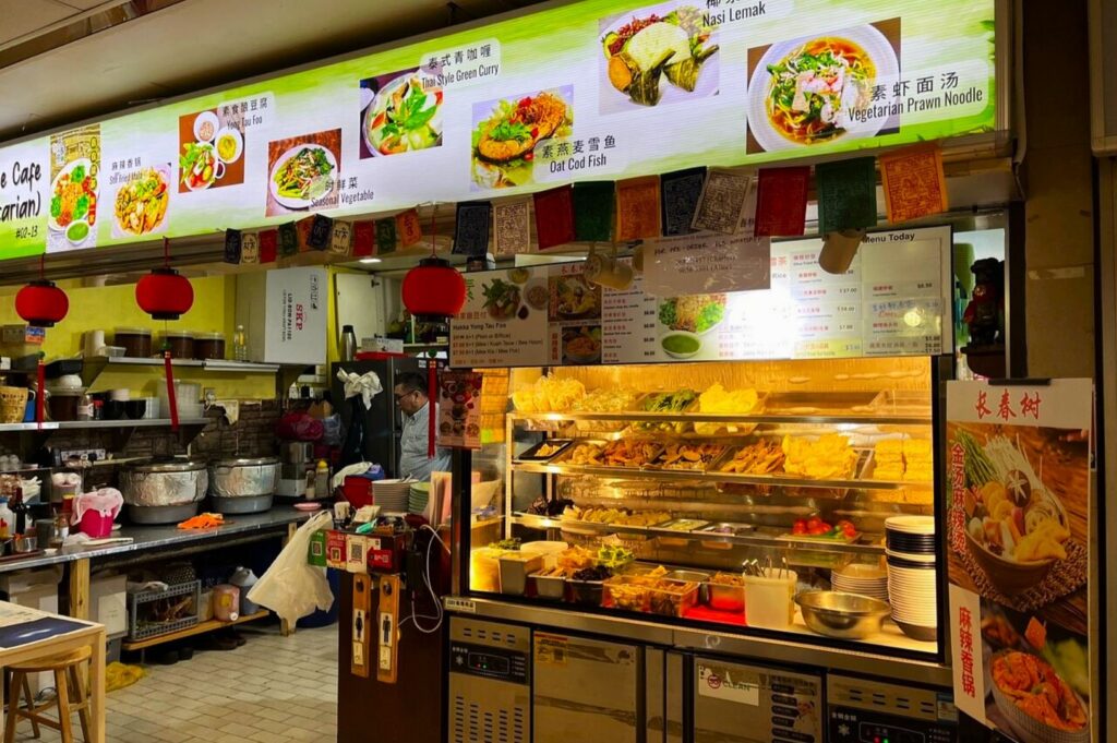 A vibrant food stall showcasing vegetarian dishes with illuminated menu signs above, colorful hanging flags, and a warm, inviting atmosphere. Kitchen tools and ingredients are visible.