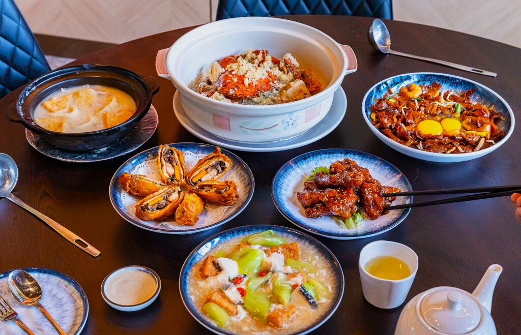 A dining table with diverse Asian dishes, including a hot pot, fried crab, egg rolls, stir-fry, and melon soup, creating a vibrant and appetizing scene.