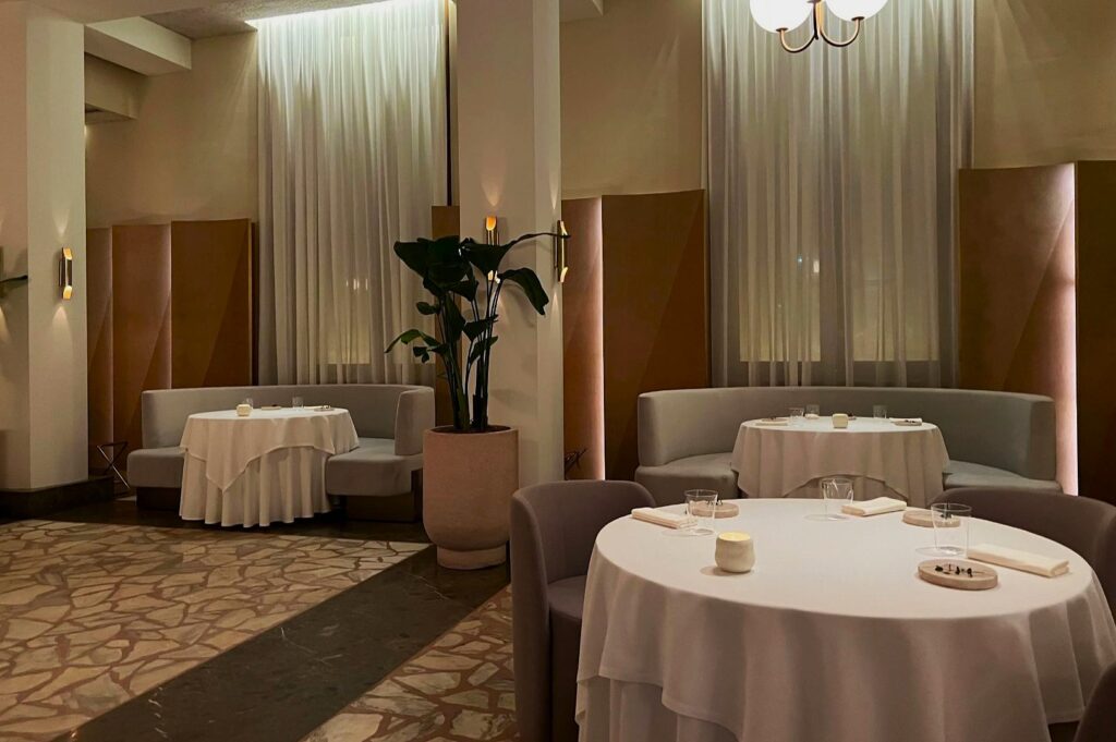 Elegant restaurant interior with round tables draped in white cloths, set for fine dining. Soft lighting, potted plant, and serene atmosphere with muted tones.