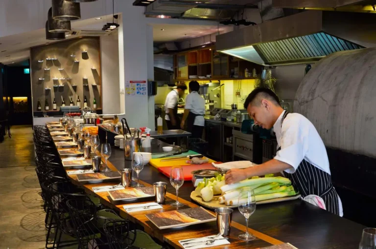 Modern kitchen with chefs. One chef prepares vegetables on a counter covered with neatly set tables and glasses, creating a busy yet inviting atmosphere.