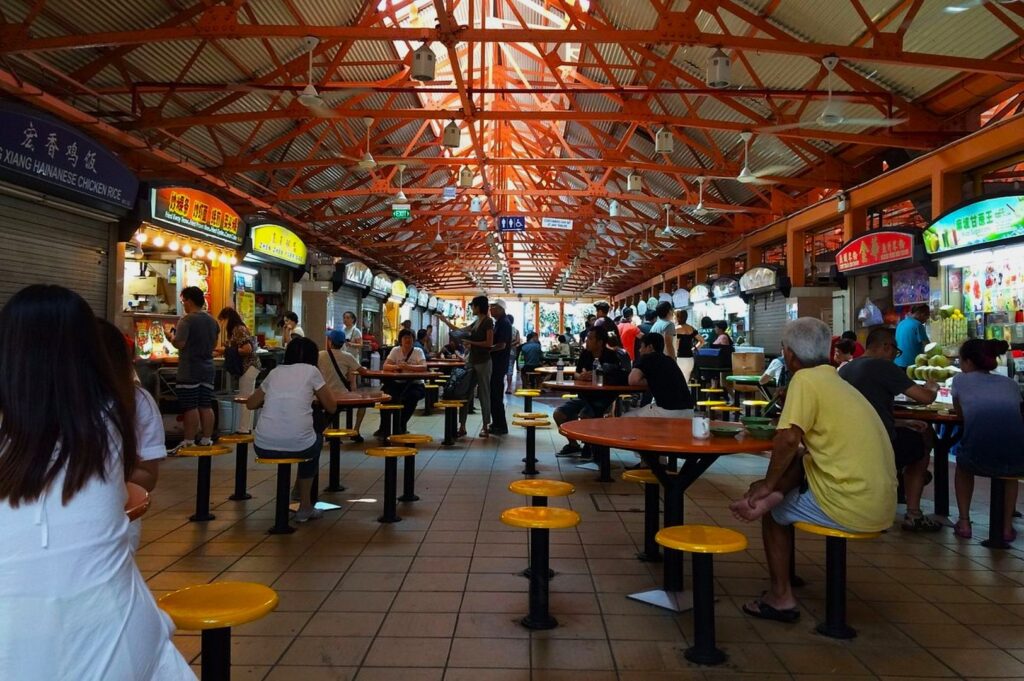 Bustling food market with red metal beams, patrons seated at round tables with yellow stools, surrounded by various food stalls. Vibrant, lively atmosphere.