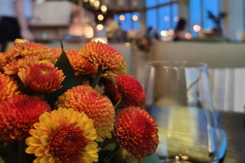 A centerpiece of vibrant orange and yellow flowers with a blurred cafe background. Soft lighting and a warm, inviting ambiance fill the scene.
