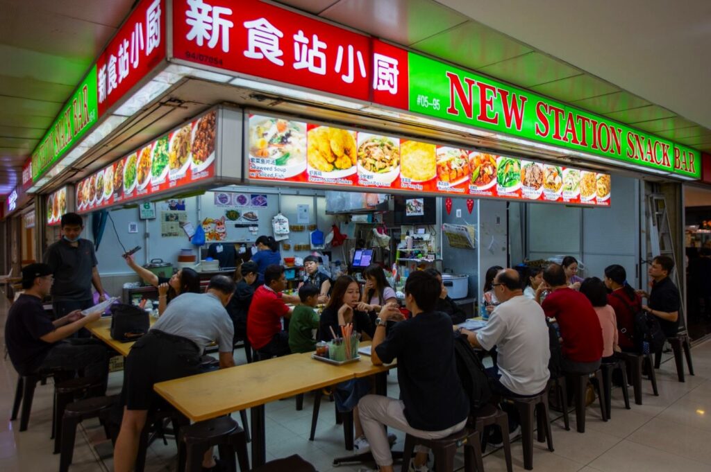 A bustling food court stall, "New Station Snack Bar," features colorful menu images. Patrons dine at simple tables, creating a lively and communal atmosphere.