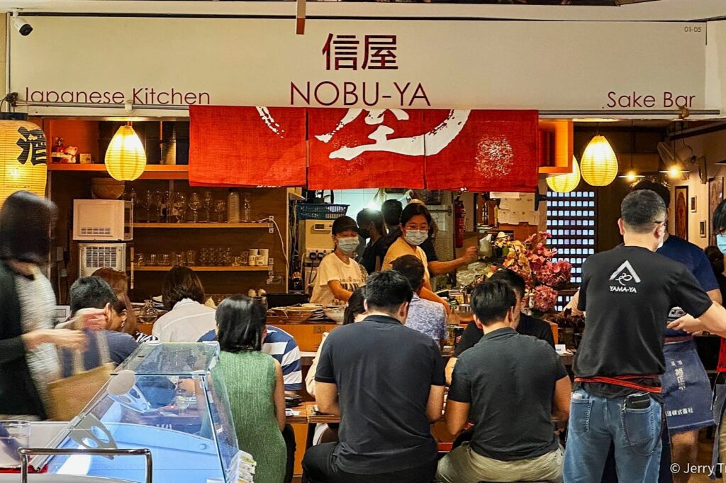 Bustling Japanese kitchen, "Nobu-Ya," with patrons seated at a counter. Warm lighting, red decor, and staff wearing masks, creating a lively, cozy atmosphere.