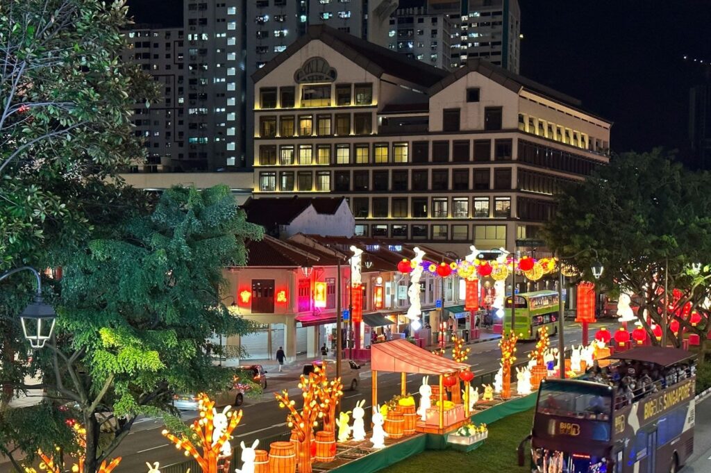 Festive city street at night with colorful lantern decorations, rabbits, and trees. Bright lights illuminate traditional buildings and a passing open-top bus.