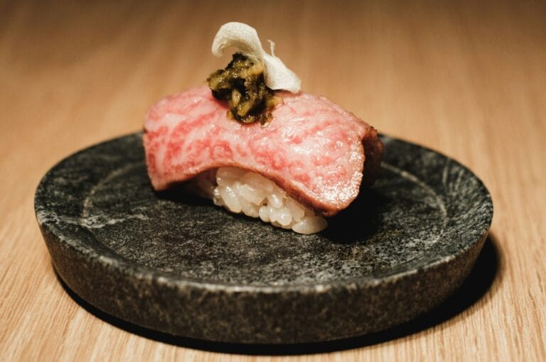 Close-up of delicate sushi on a black stone plate. The sushi features marbled pink fish, white rice, and a green topping, set against a wooden table.