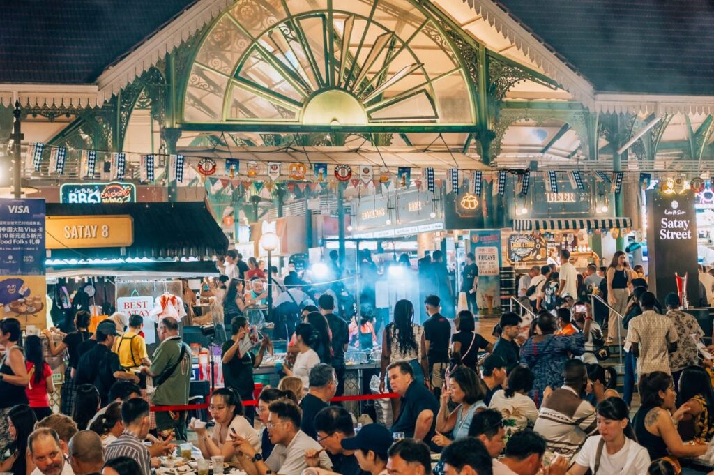 Bustling night market scene with diverse crowd dining and exploring food stalls. Bright lights and decorative banners create a lively, festive atmosphere.