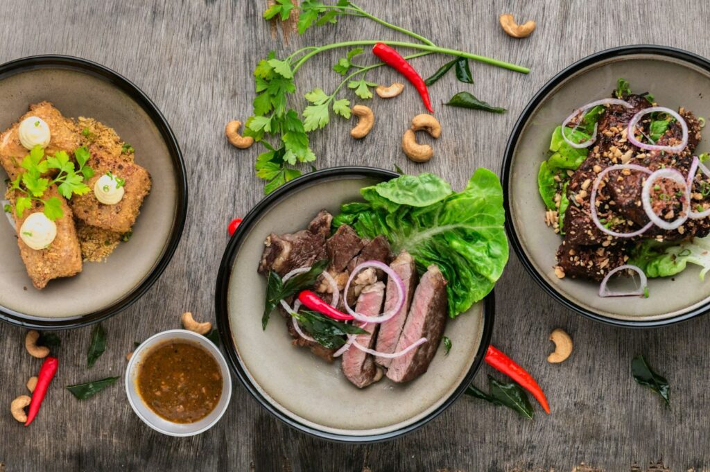 Three dishes on a wooden table: crispy rolls with herbs, sliced steak with lettuce and onion, and seasoned meat with cashew and onion. Surrounding garnishes include red chilies, coriander, and cashews.