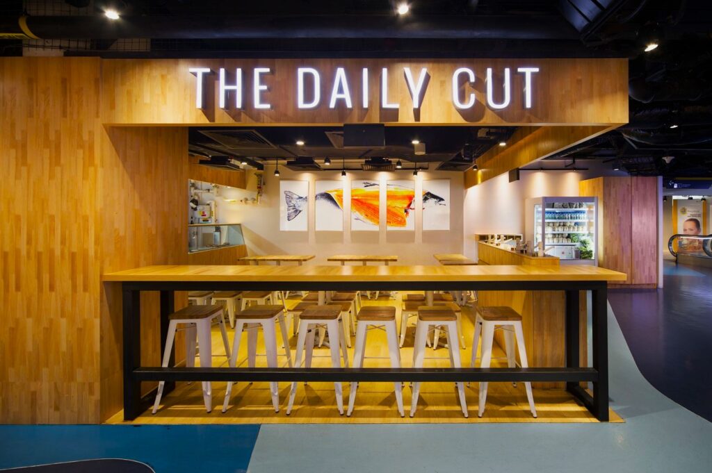Modern eatery "The Daily Cut" with wood decor, high stools, and bright lighting. Features vibrant fish art, open kitchen, and inviting atmosphere.