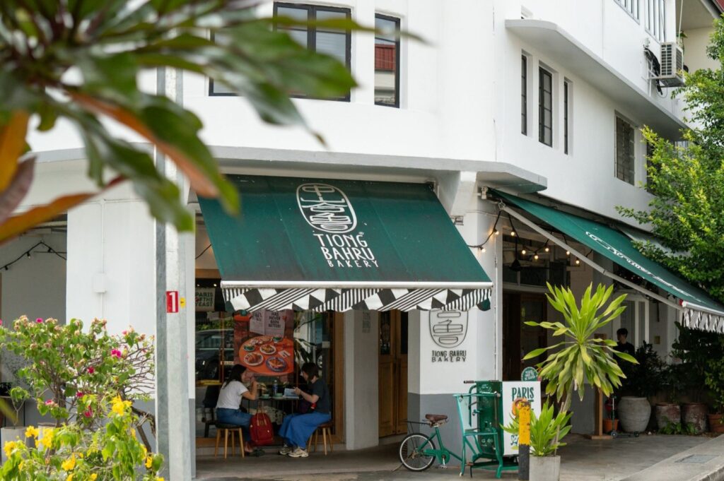 Street view of a cozy bakery with green awnings displaying "Tiong Bahru Bakery." Patrons sit at outdoor tables, surrounded by lush plants and bicycles.