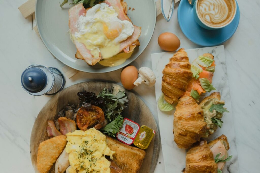 A delicious brunch spread featuring eggs Benedict, croissants with ham and salad, scrambled eggs, hash browns, a cappuccino, and assorted condiments.