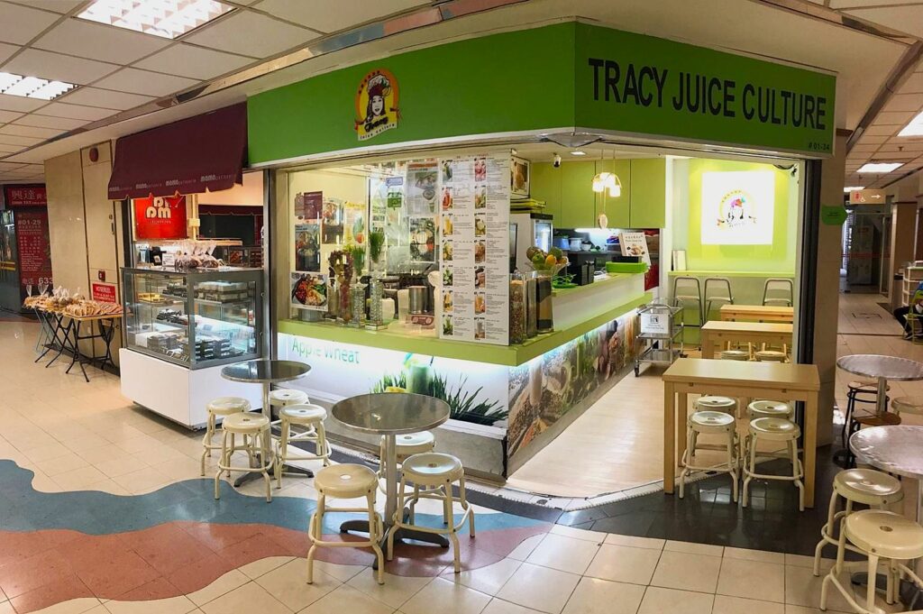 Bright juice bar with green signage reading "Tracy Juice Culture," features a cozy interior, round tables with stools, menu display, and vibrant decor.