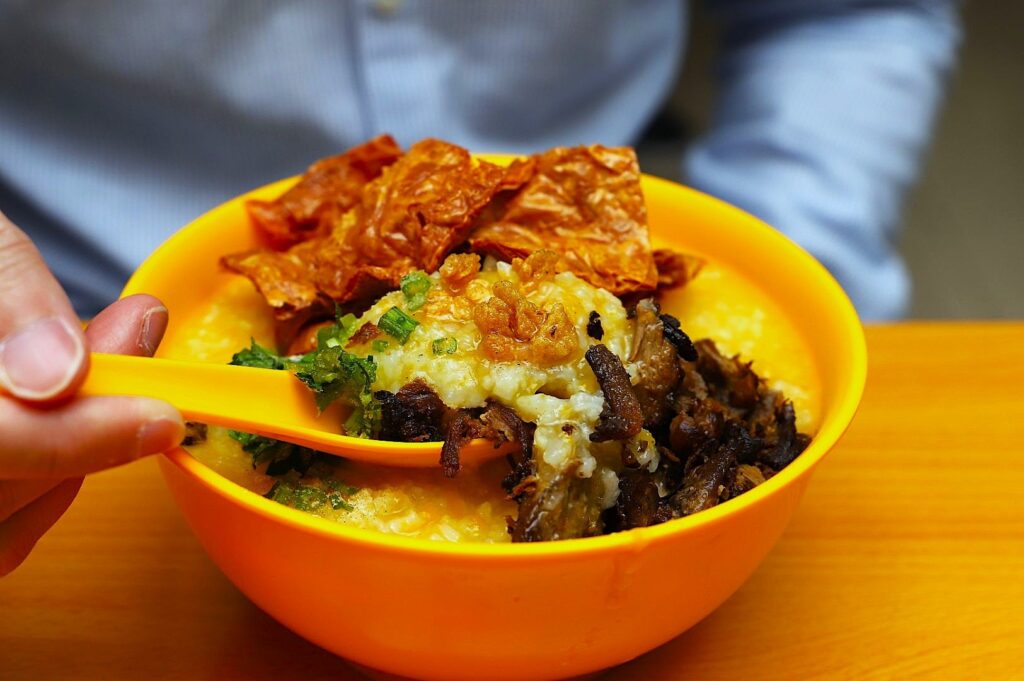 A vibrant yellow bowl filled with creamy porridge, topped with crispy fried shallots, herbs, and crunchy chips. A hand holds a matching yellow spoon.