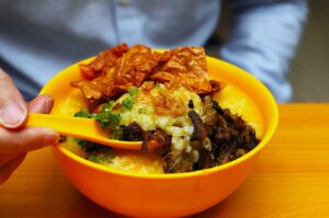 A vibrant yellow bowl filled with creamy porridge, topped with crispy fried shallots, herbs, and crunchy chips. A hand holds a matching yellow spoon.