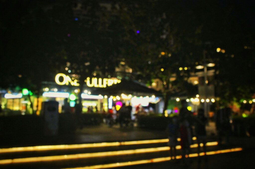 Blurred night scene of a lit plaza with glowing storefronts, people silhouetted against softly illuminated steps. The ambiance is lively yet tranquil.