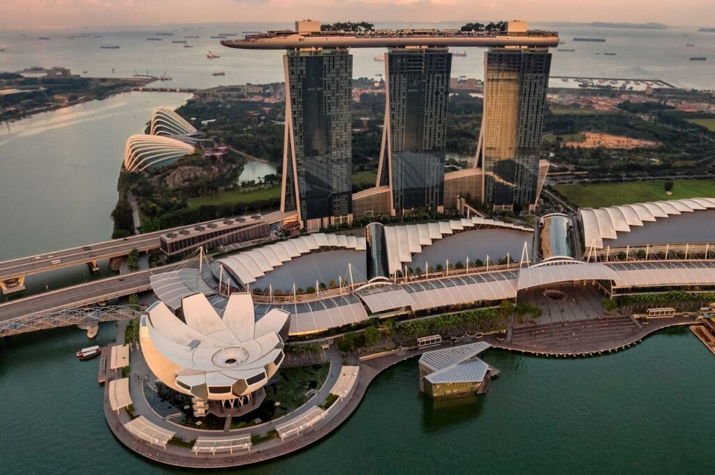 Aerial view of Marina Bay Sands at sunset in Singapore, showcasing the iconic three-tower structure with a rooftop park. Nearby, the lotus-shaped ArtScience Museum and surrounding waters are visible, with a calm and serene atmosphere.