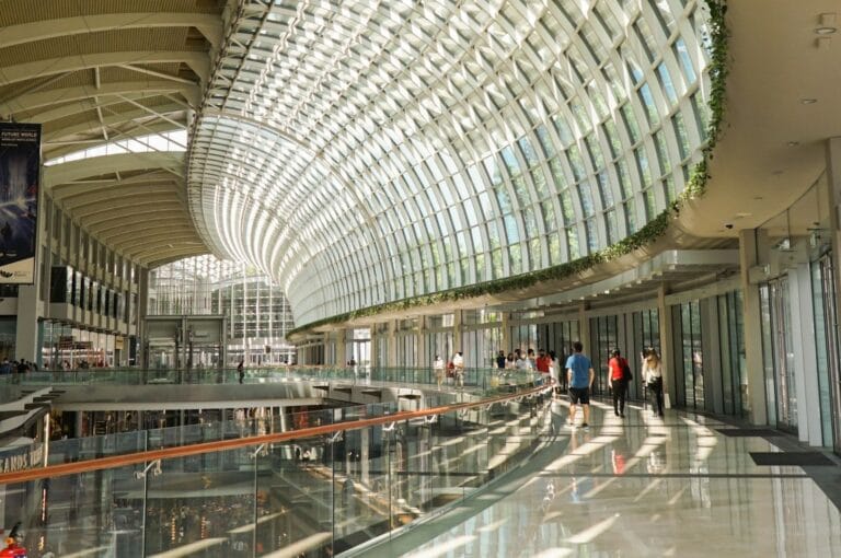 Spacious modern mall interior with arched glass ceiling, sunlit, and lined with plants. People walk along shiny floors, creating an airy, bustling atmosphere.