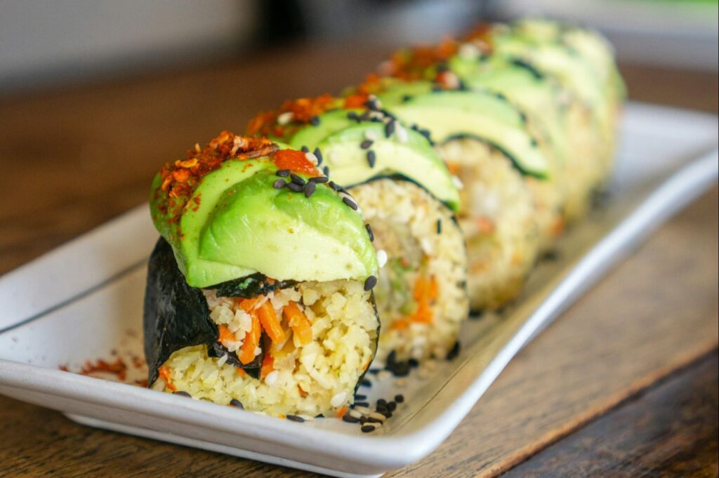 Close-up of sushi rolls topped with avocado slices and sesame seeds on a white plate. The rolls are filled with rice and vegetables, conveying freshness.