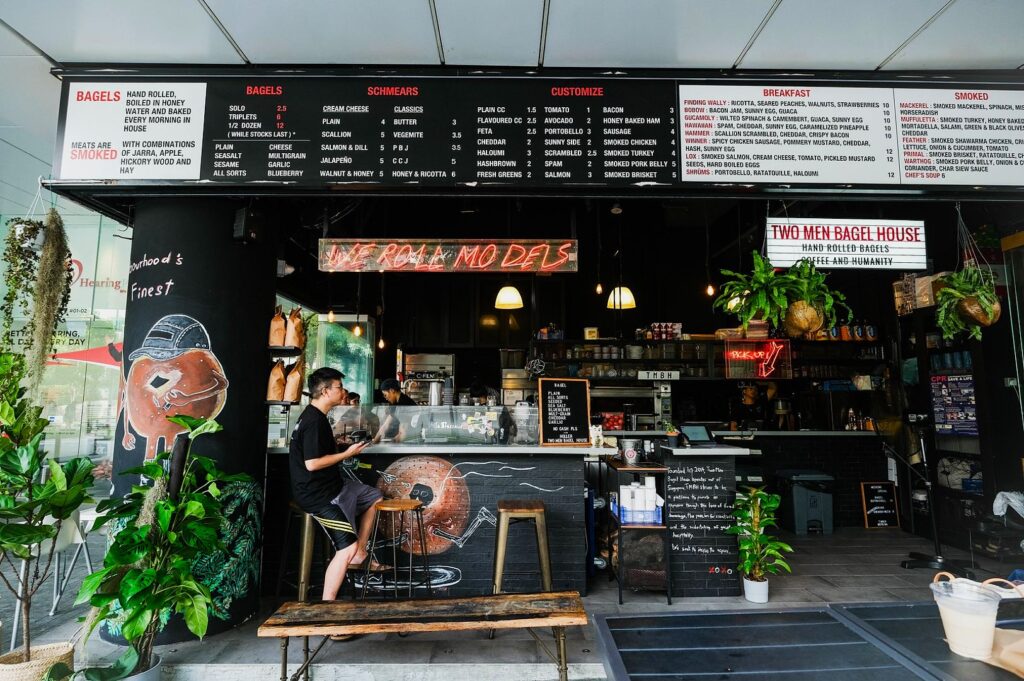 A cozy bagel shop with a blackboard menu displays various options. A person sits at the counter on a stool. Lush plants add a vibrant touch.