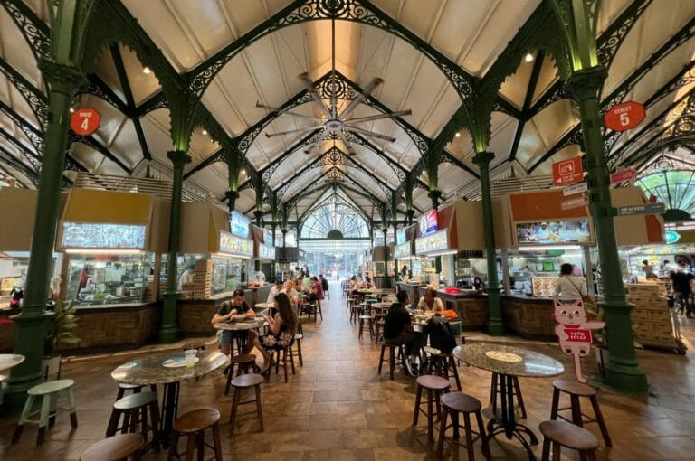 A bustling indoor food court with ornate green metal arches and a high ceiling. People are seated at round tables enjoying meals, creating a lively atmosphere.