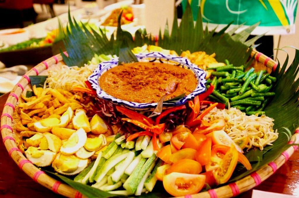 A vibrant platter of Gado-Gado is presented with a central brown peanut sauce surrounded by a colorful array of sliced vegetables and boiled eggs, all garnished with banana leaves.
