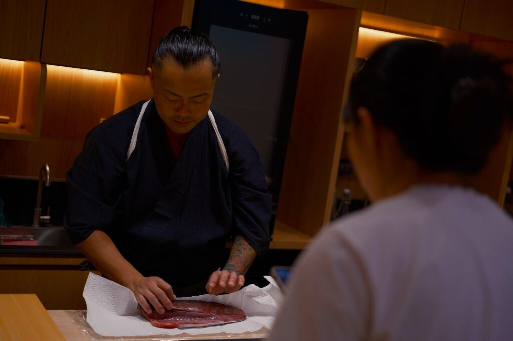 Chef in a dark outfit focuses on preparing a large piece of fish on a counter. A person in the foreground watches. The setting is warm and cozy.