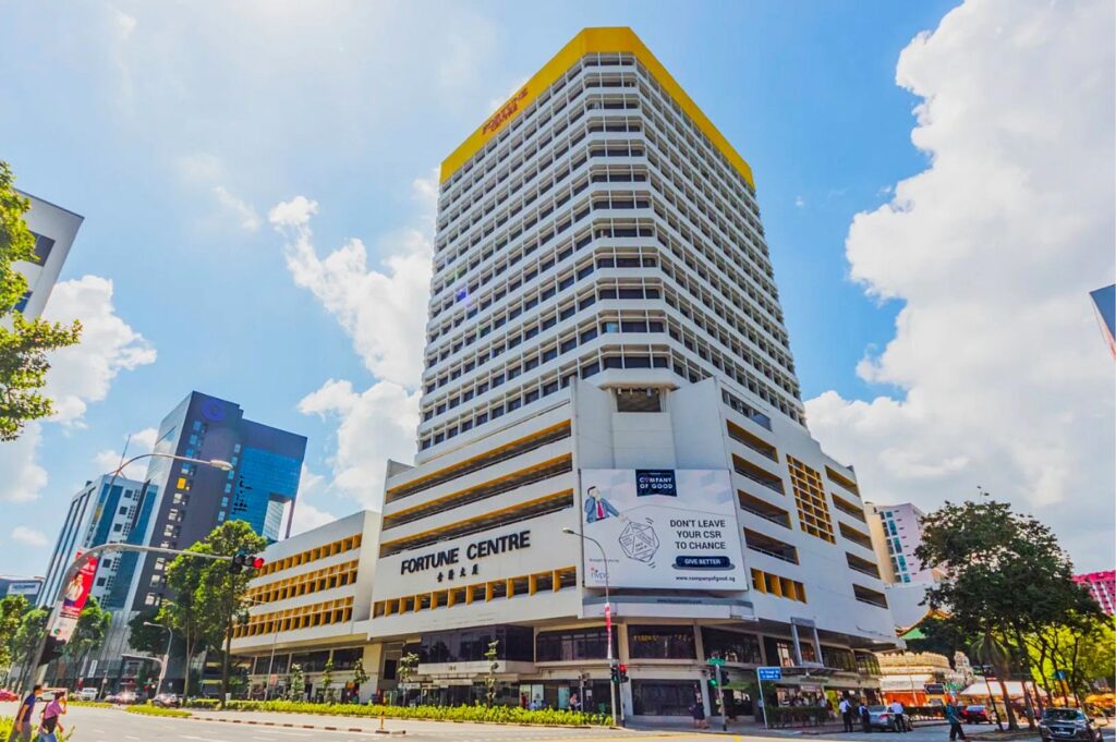 A tall, modern building labeled "Fortune Centre" with a yellow and white facade under a vibrant blue sky. The scene conveys a bustling urban atmosphere.
