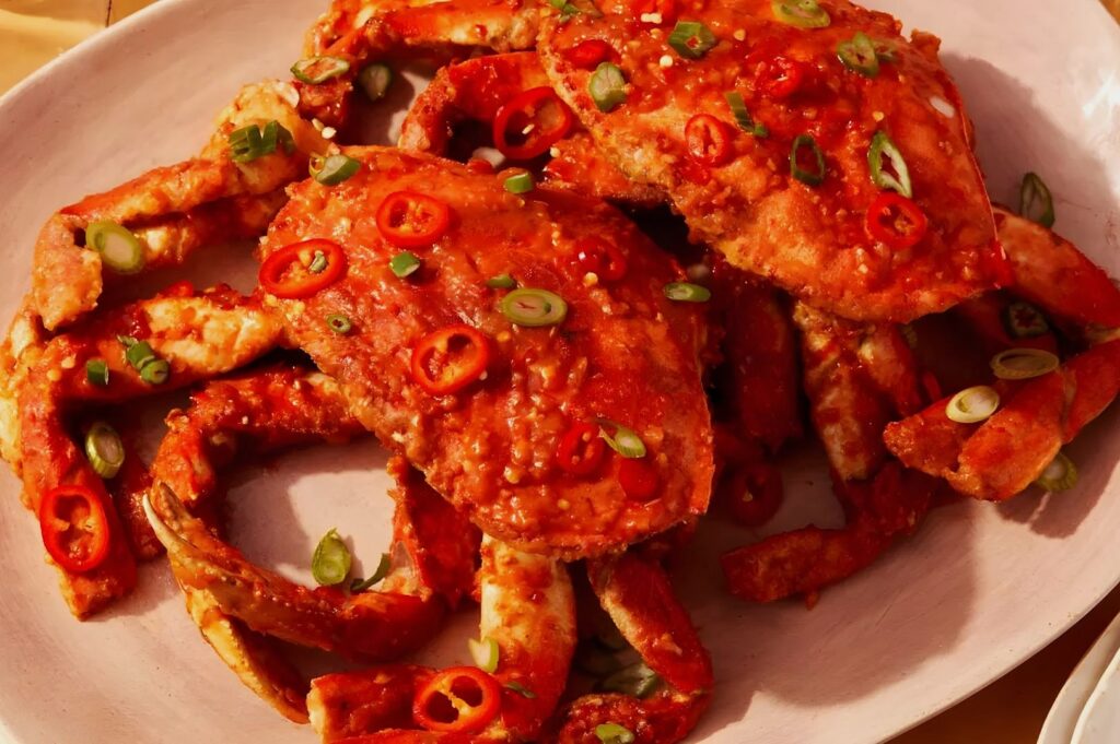 Spicy Asian-style crab dish with vibrant red sauce, garnished with sliced red chilies and green onions, served on a white plate, evoking a savory and spicy aroma.