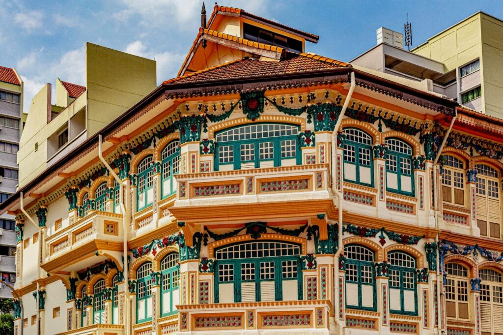 A vibrant Peranakan-style building with ornate, multicolored facades and green shutters. It stands under a clear blue sky, surrounded by modern buildings.